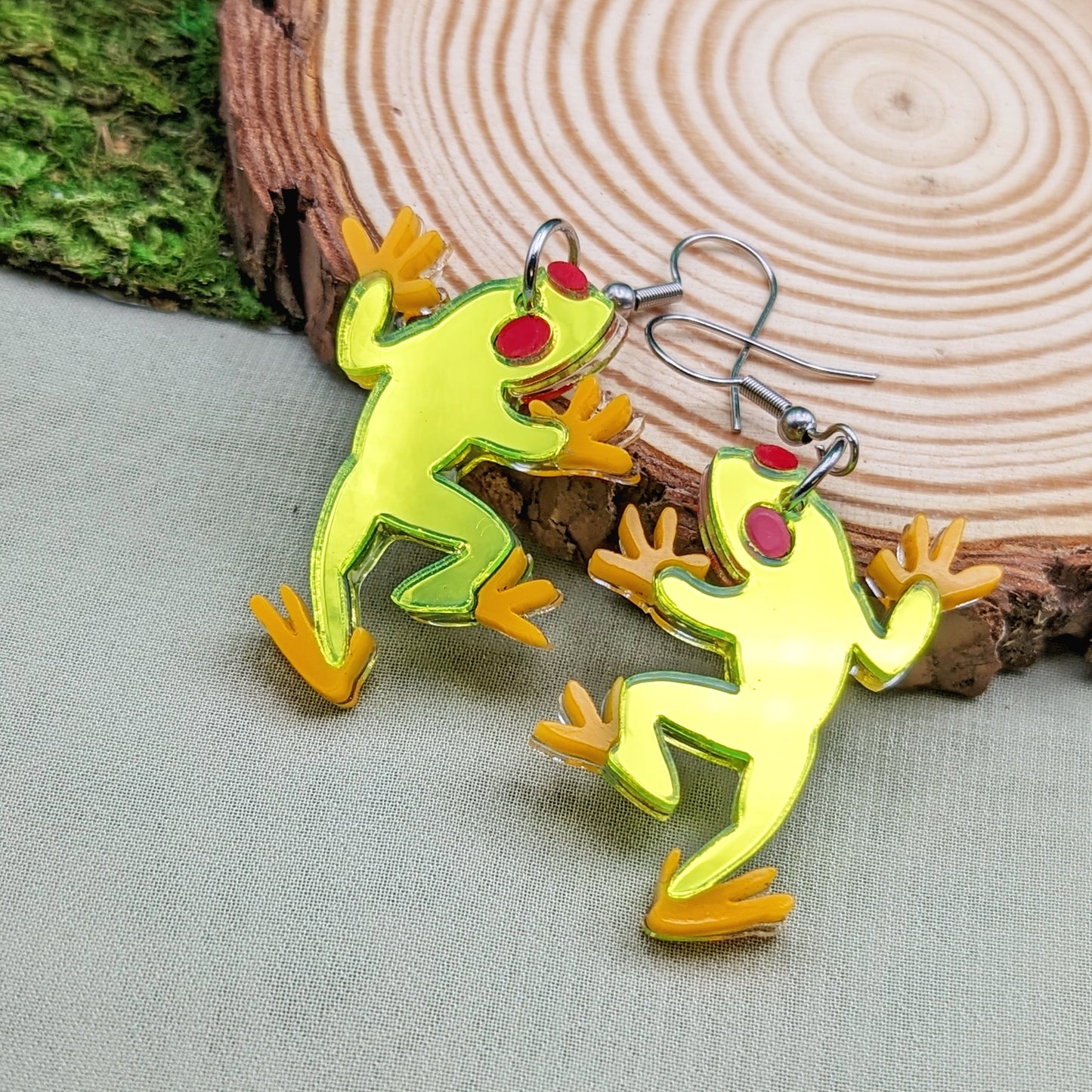 Neon green tree frog earrings with red eyes and orange feet on a wood slice