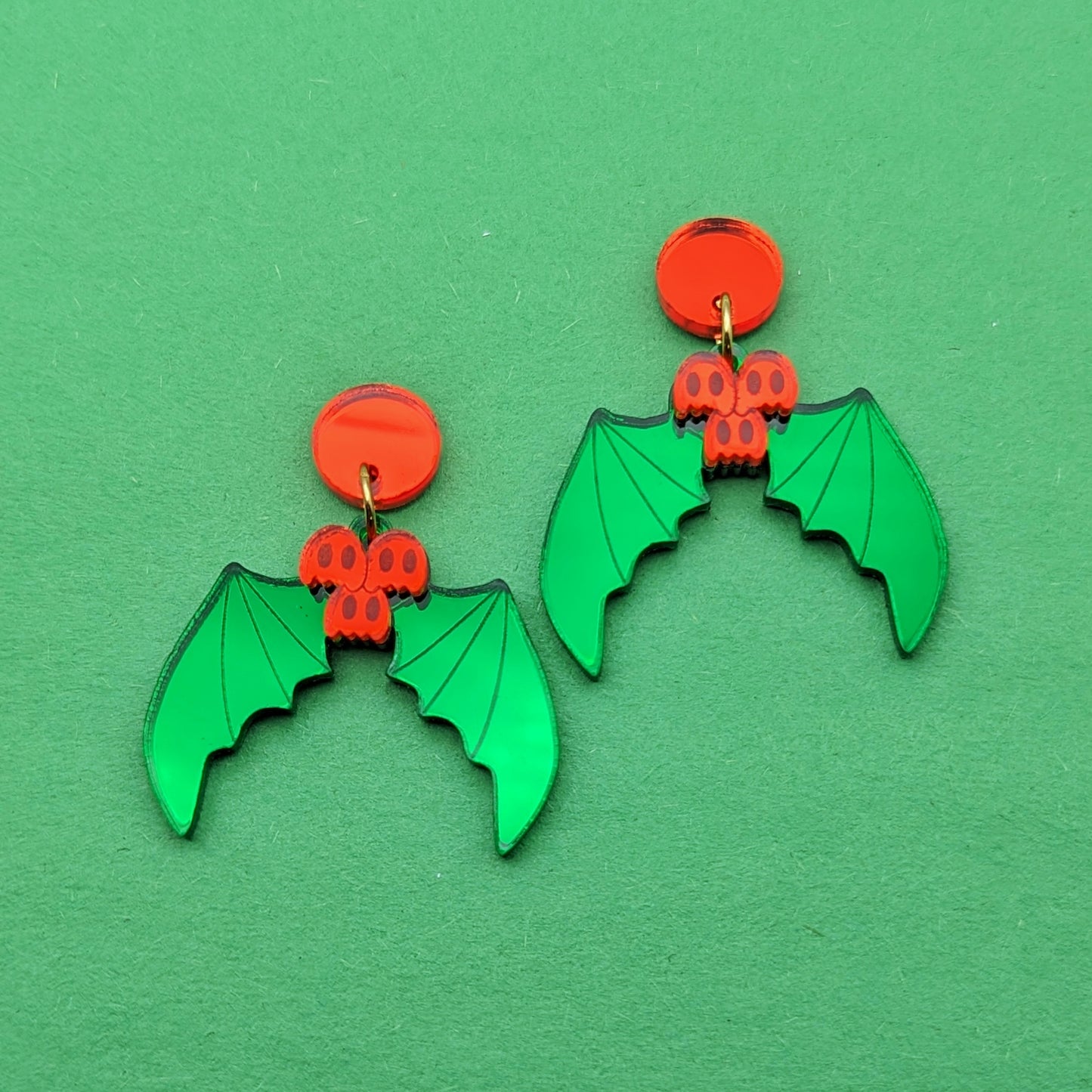 Holly shaped earrings with green leaves shaped like bat wings and red berries shaped like ghosts. They have gold-toned jump rings and red circular stud posts.