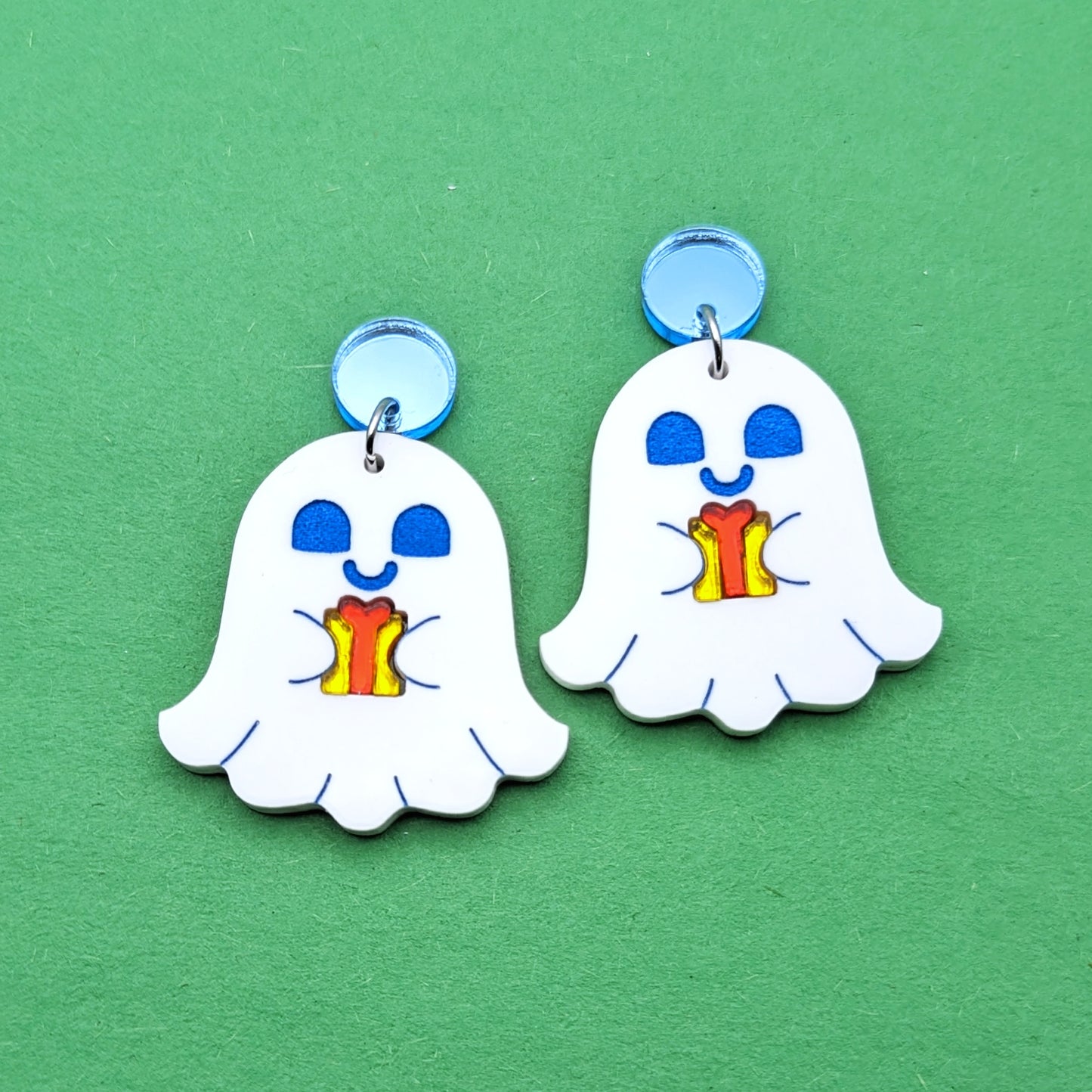 White ghost shaped earrings with blue engraved faces. They are holding yellow presents with red ribbon. Stainless steel jump rings connect to light blue circular stud posts.