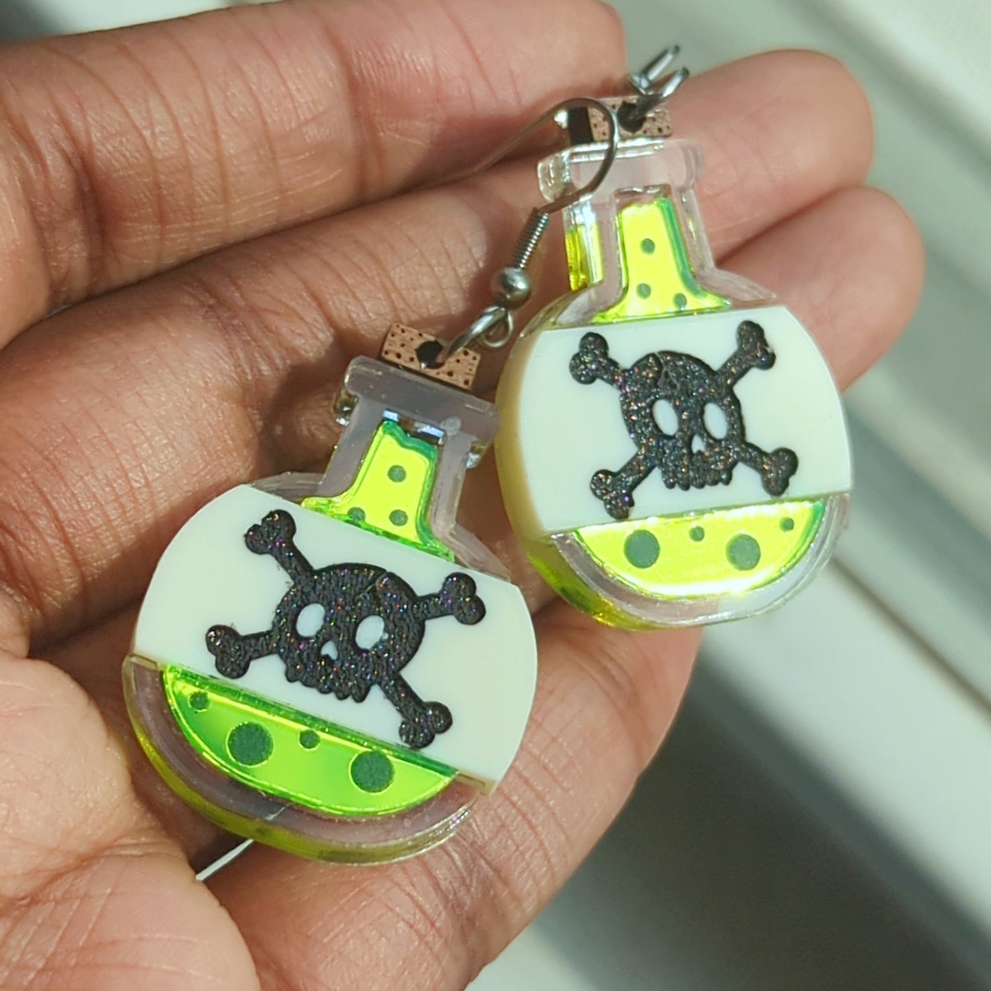 Poison bottle earrings with neon green liquid in a clear bottle with a beige label bearing a skull and crossbones with a cork on top