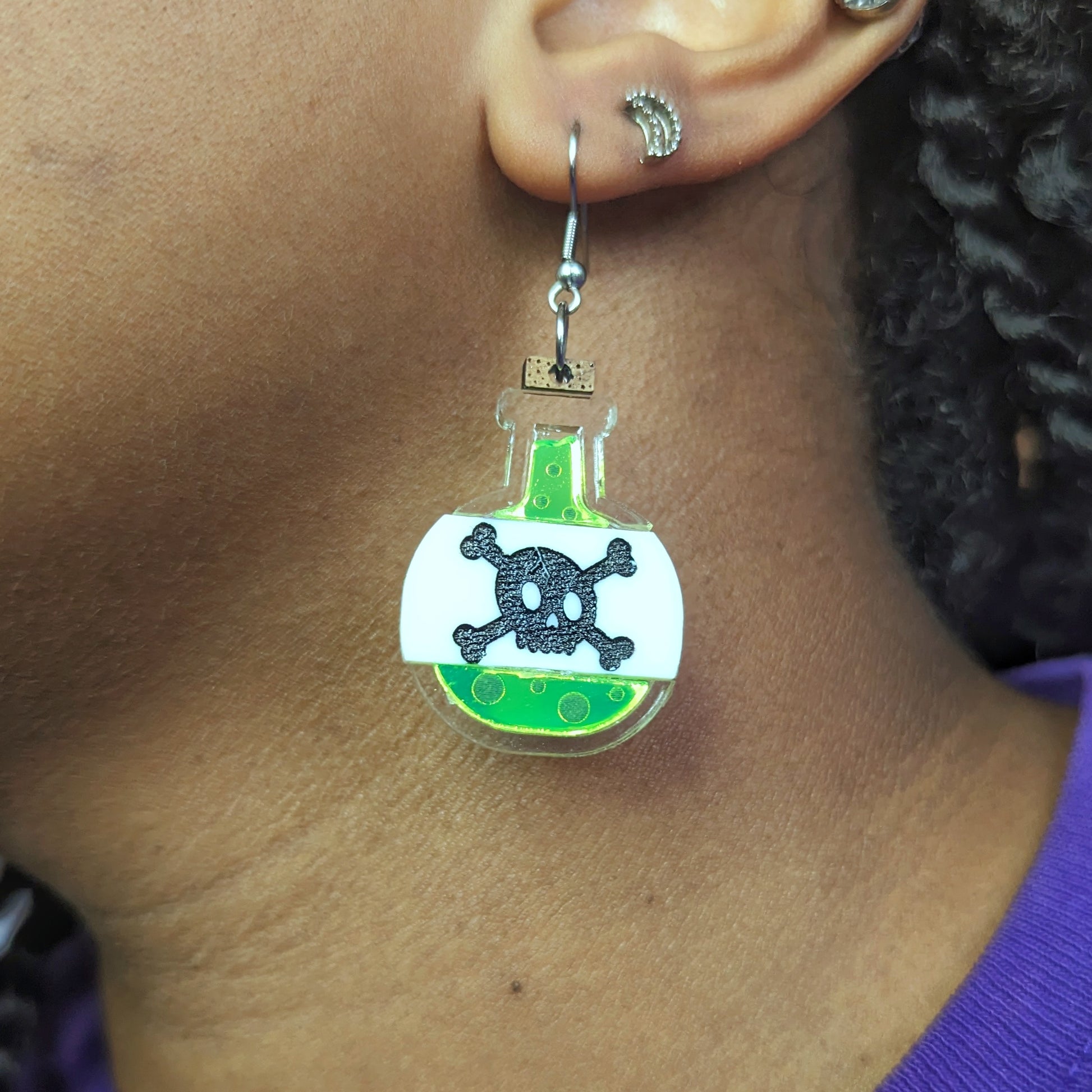 Poison bottle earrings with neon green liquid in a clear bottle with a beige label bearing a skull and crossbones with a cork on top