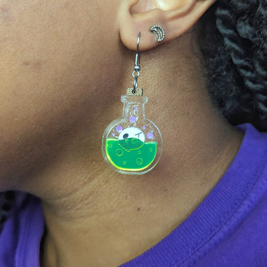 Skull potion bottle earrings with a white skill floating in neon green liquid, with purple bubbles, in a clear bottle with a cork on top