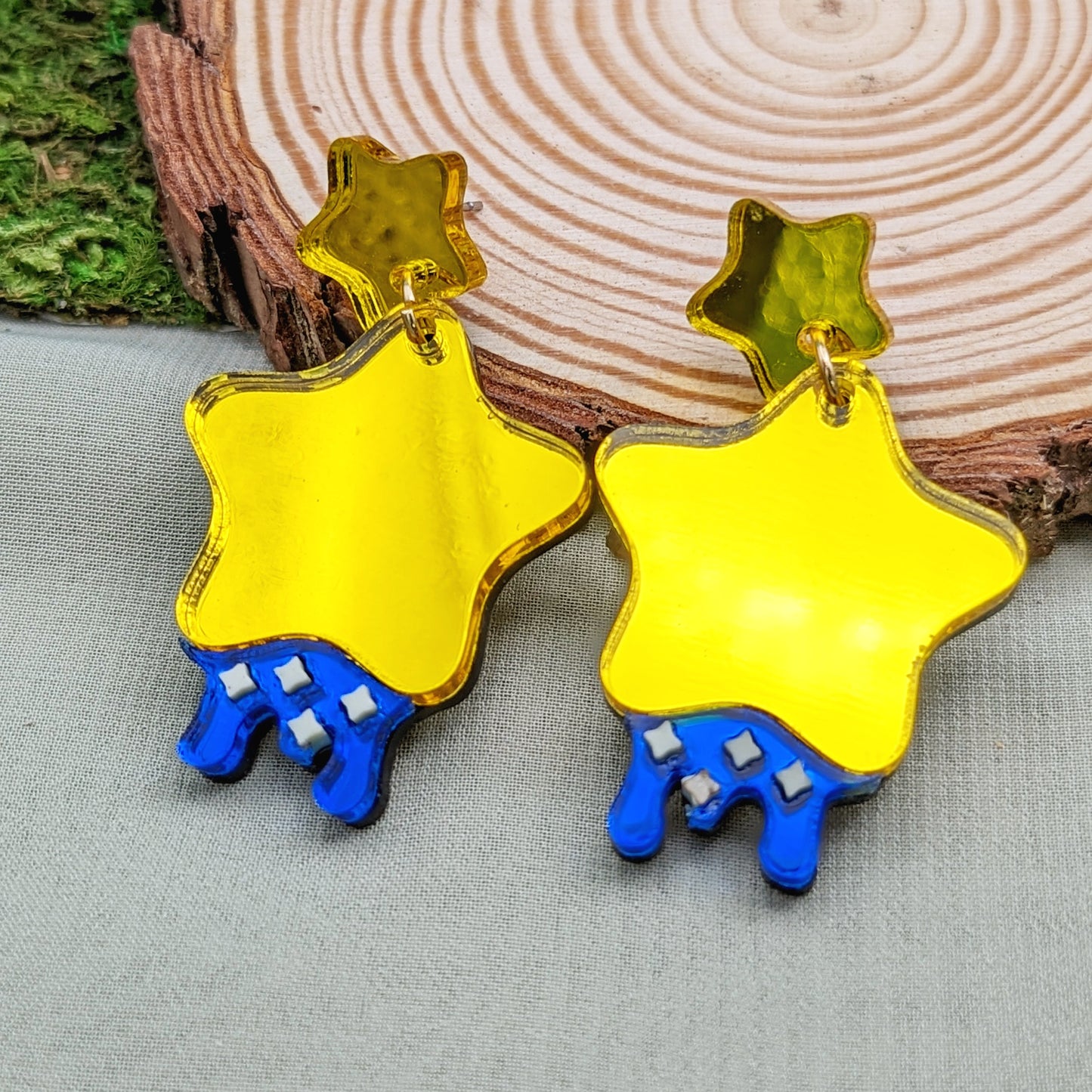 yellow star shaped dangle earrings with blue dripping below and small pastel yellow stars with yellow star stud posts on a wood slice.