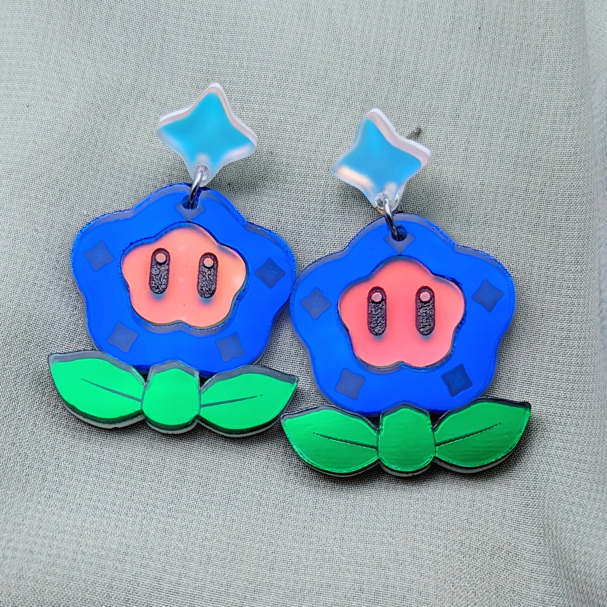 Magical flower earrings with blue petals engraved with diamonds, green leaves, and a matte iridescent face with engraved black eyes.