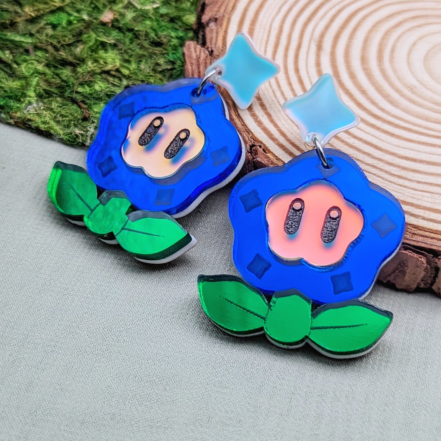 Magical flower earrings with blue petals engraved with diamonds, green leaves, and a matte iridescent face with engraved black eyes on a wood slice.