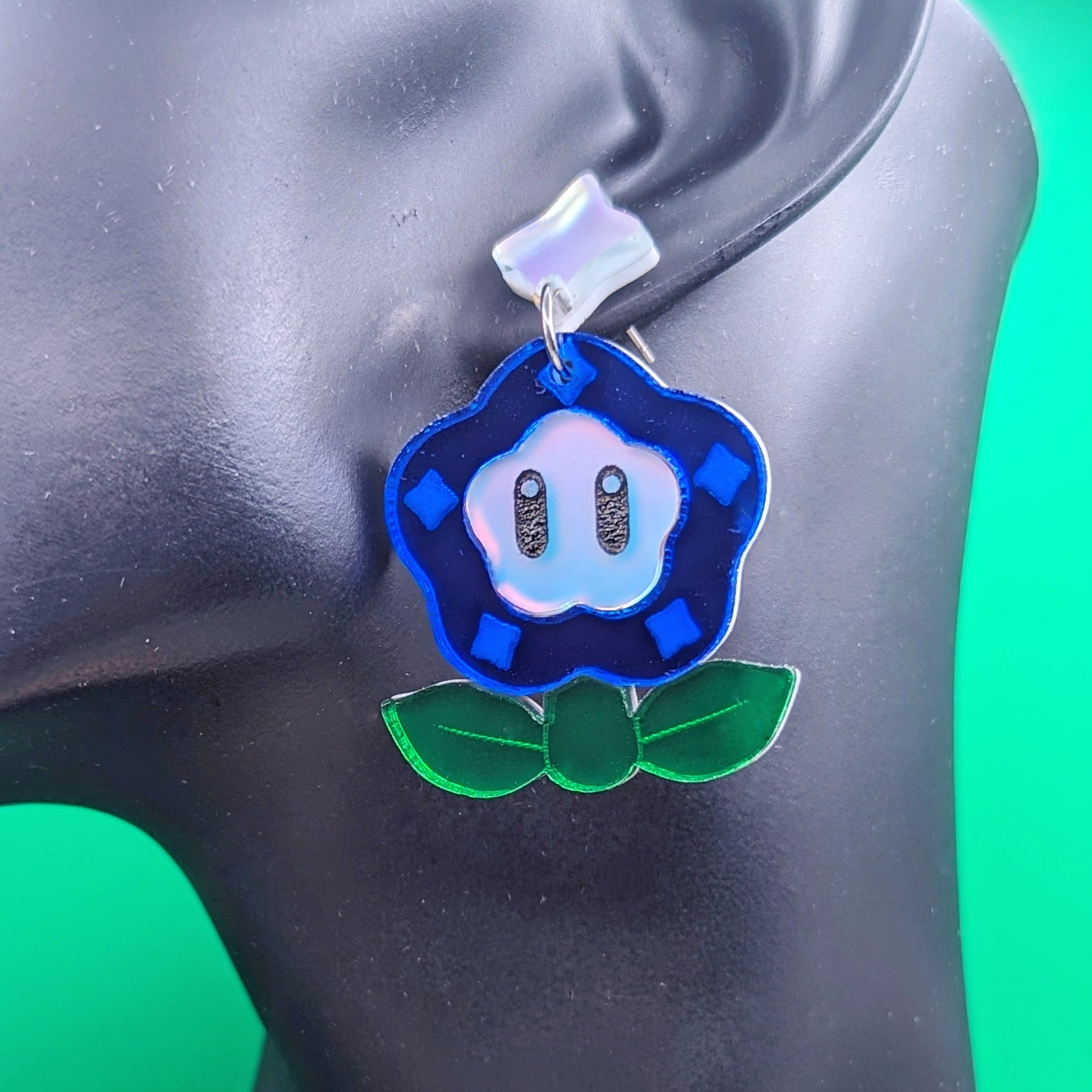 Magical flower earrings with blue petals engraved with diamonds, green leaves, and a matte iridescent face with engraved black eyes on a model ear.