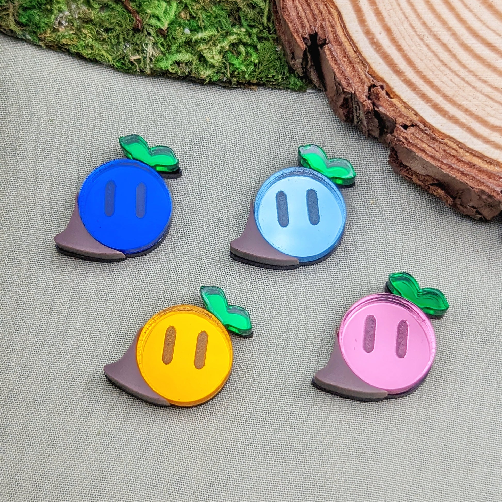 Magical seed stud earrings in dark blue, light blue, orange, and pink with green sprouts and engraved eyes next to a wood slice and moss