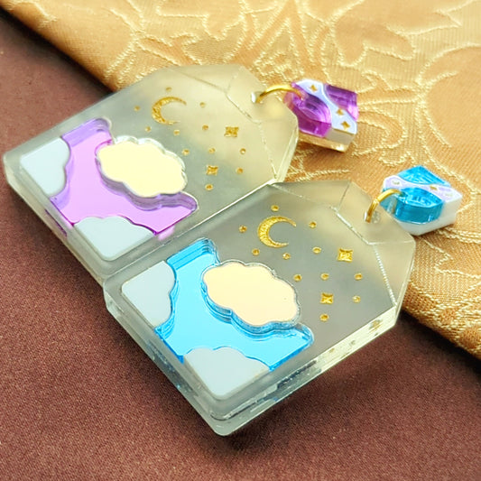 tea bag shaped earrings with blue or purple skies, white and iridescent clouds, and engraved gold moon and stars.