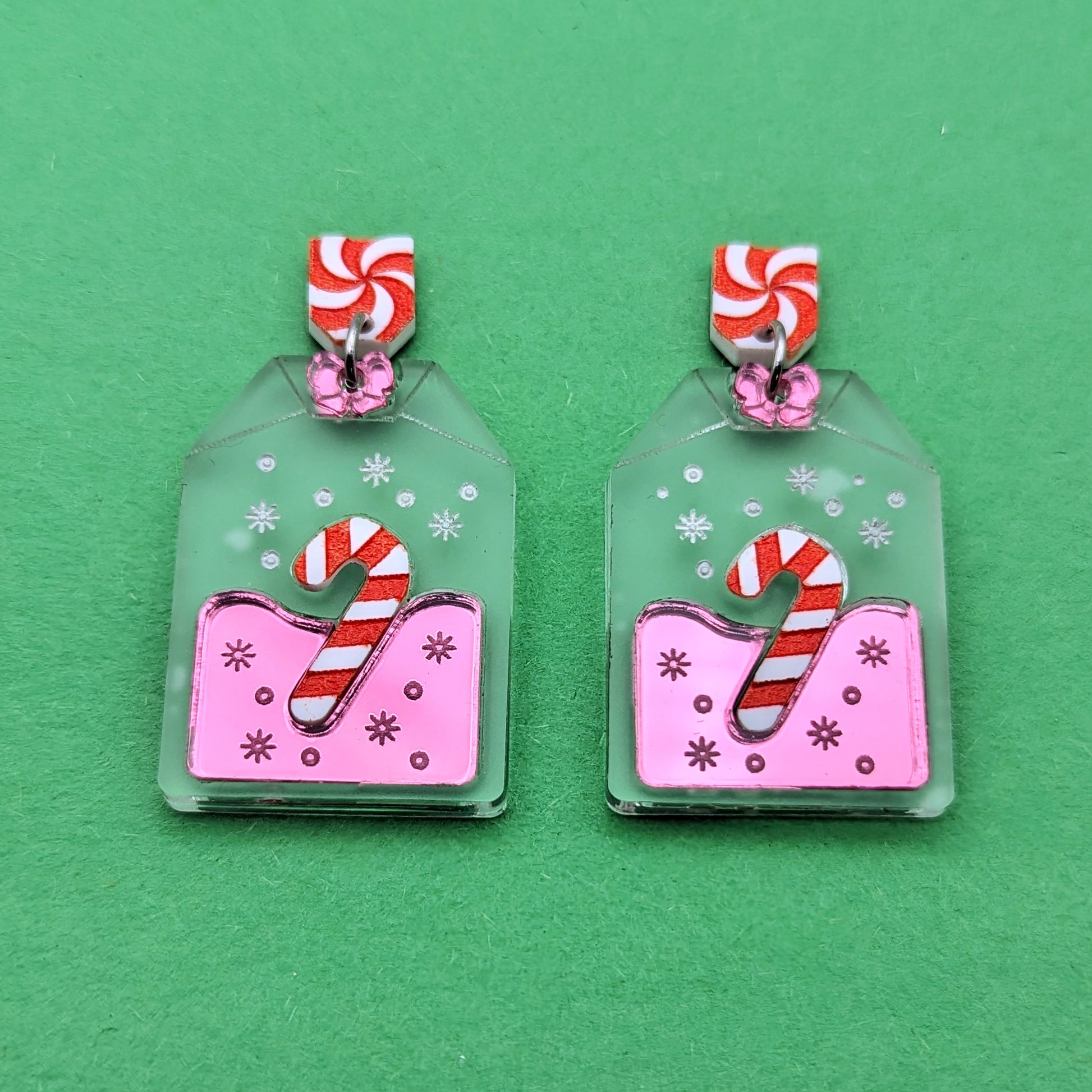 Frosted clear tea bag shaped earrings with a red and white striped candy cane in the center, surrounded by pink. There are white engraved snowflake accents and a pink bow on top. Stainless steel jump rings connect to red and white swirled pentagon stud posts.