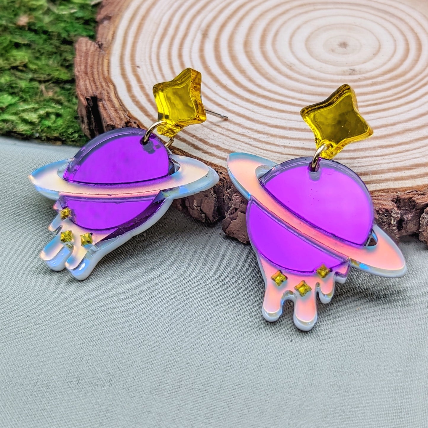 purple planet shaped dangle earrings with matte iridescent planetary rings, iridescent dripping below and small yellow stars with yellow star stud posts on a wood slice