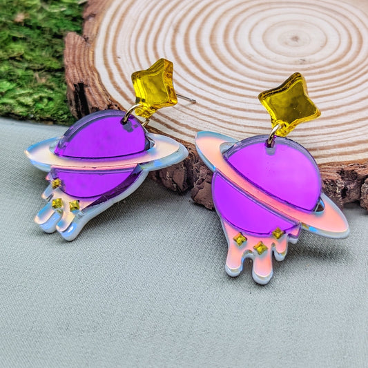 purple planet shaped dangle earrings with matte iridescent planetary rings, iridescent dripping below and small yellow stars with yellow star stud posts on a wood slice