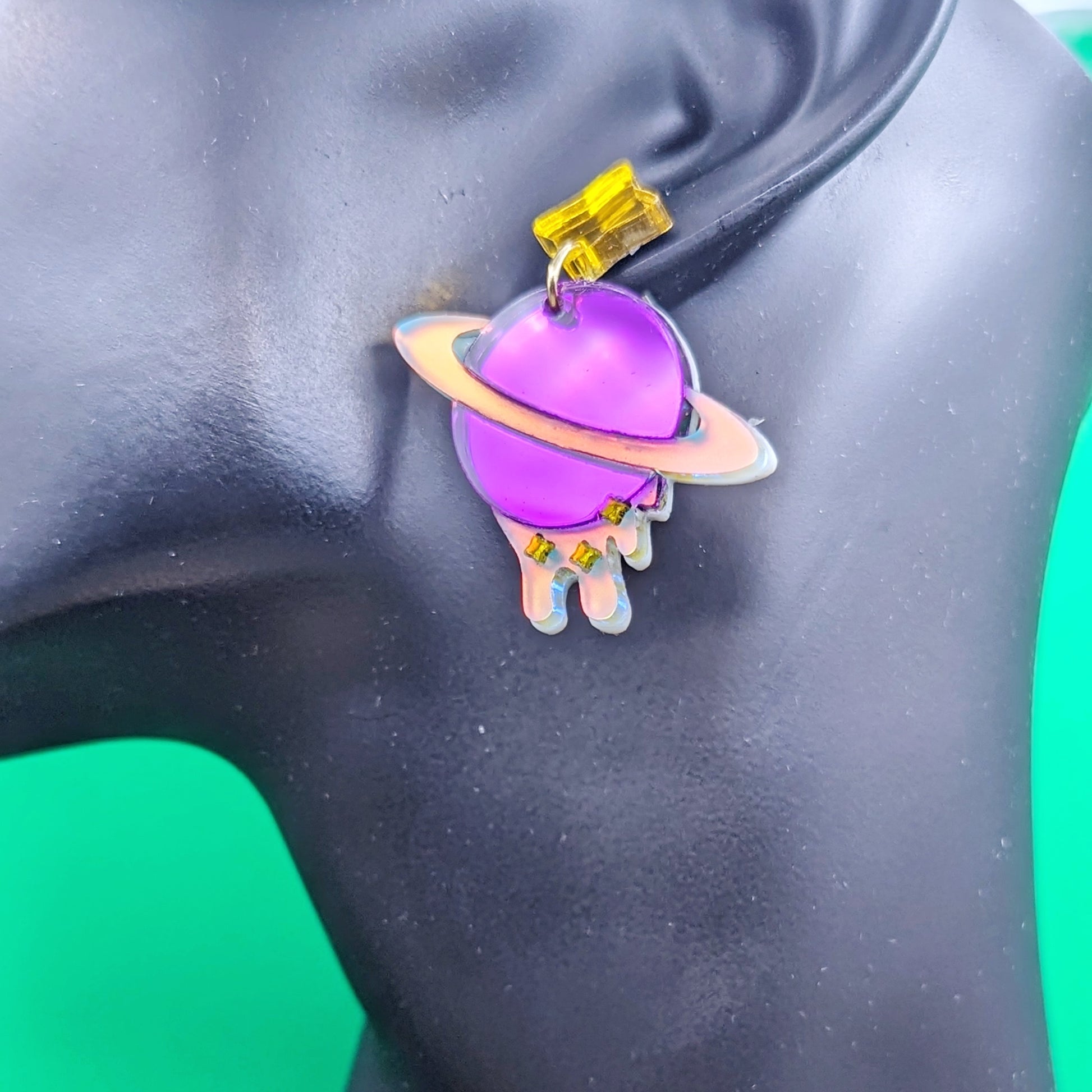 purple planet shaped dangle earrings with matte iridescent planetary rings, iridescent dripping below and small yellow stars with yellow star stud posts on a model ear