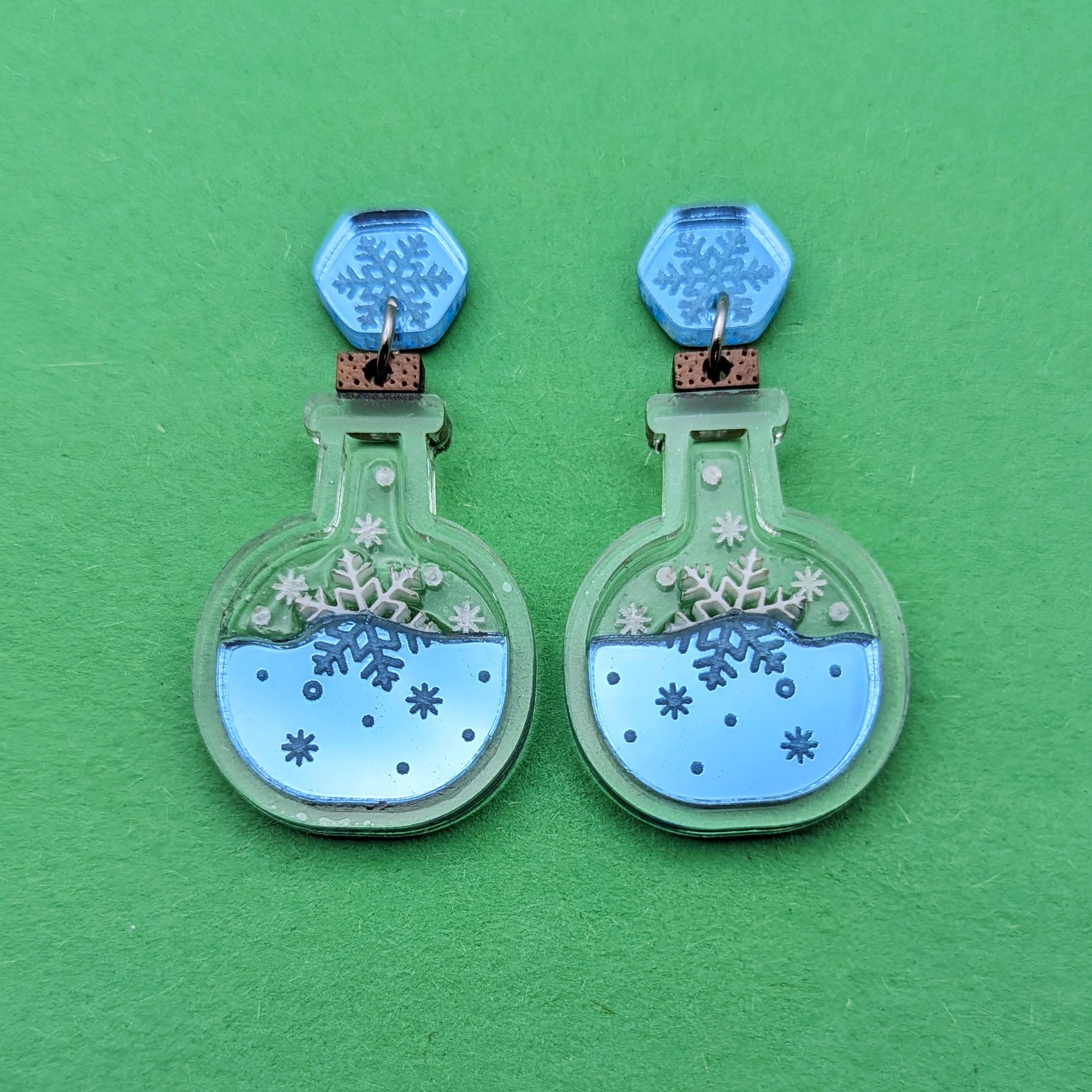 Clear bottle shaped earrings with light blue acrylic at the bottom. With white snowflake accents and topped with a wooden cork. Stainless steel jump rings connect to light blue hexagon stud posts engraved with snowflakes.