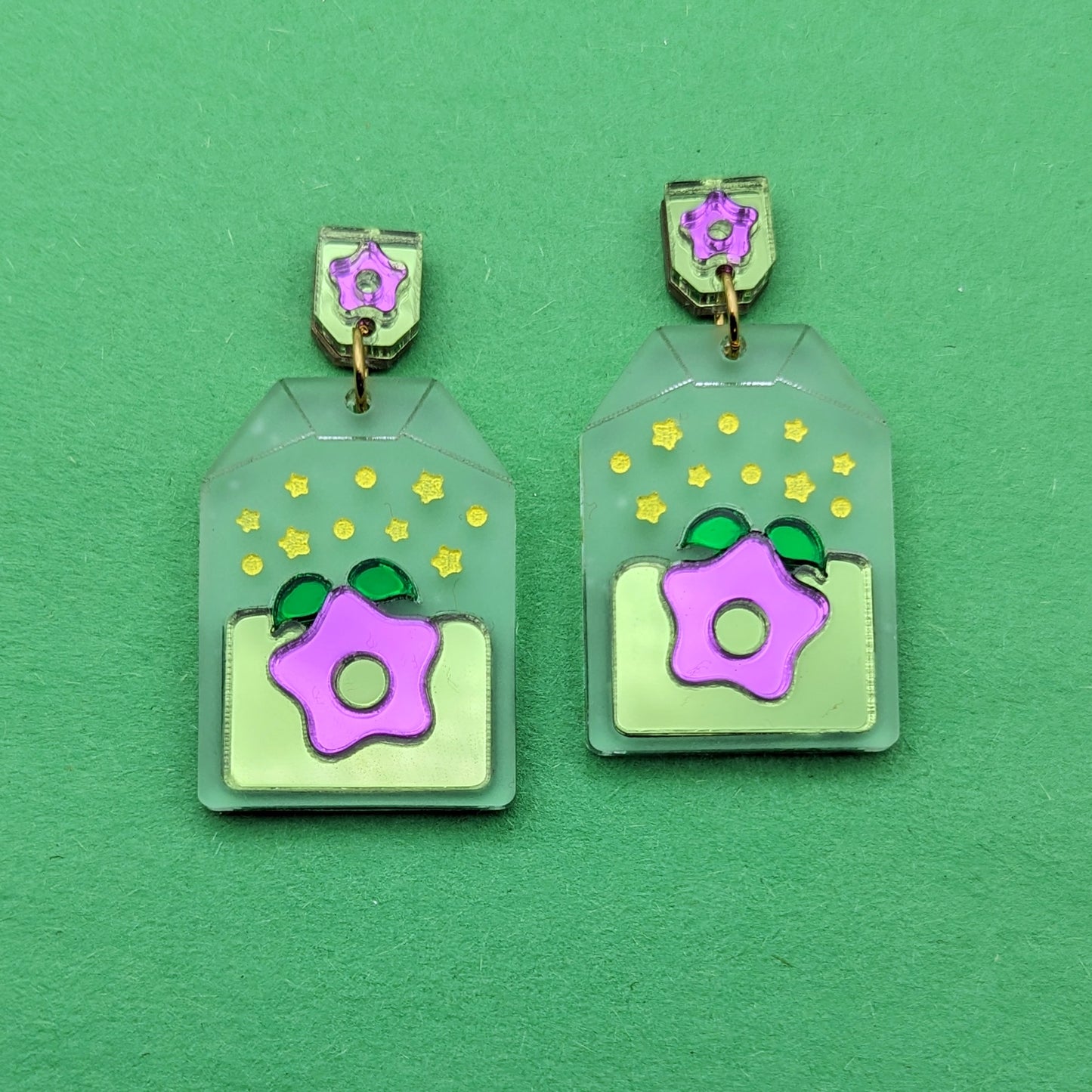 Frosted clear tea bag shaped earrings with a purple starfruit in the middle. The star has green leaves and is surrounded by light green. There are yellow star accents. Gold-toned jump rings connect to tag shaped stud posts that are green with a purple star.