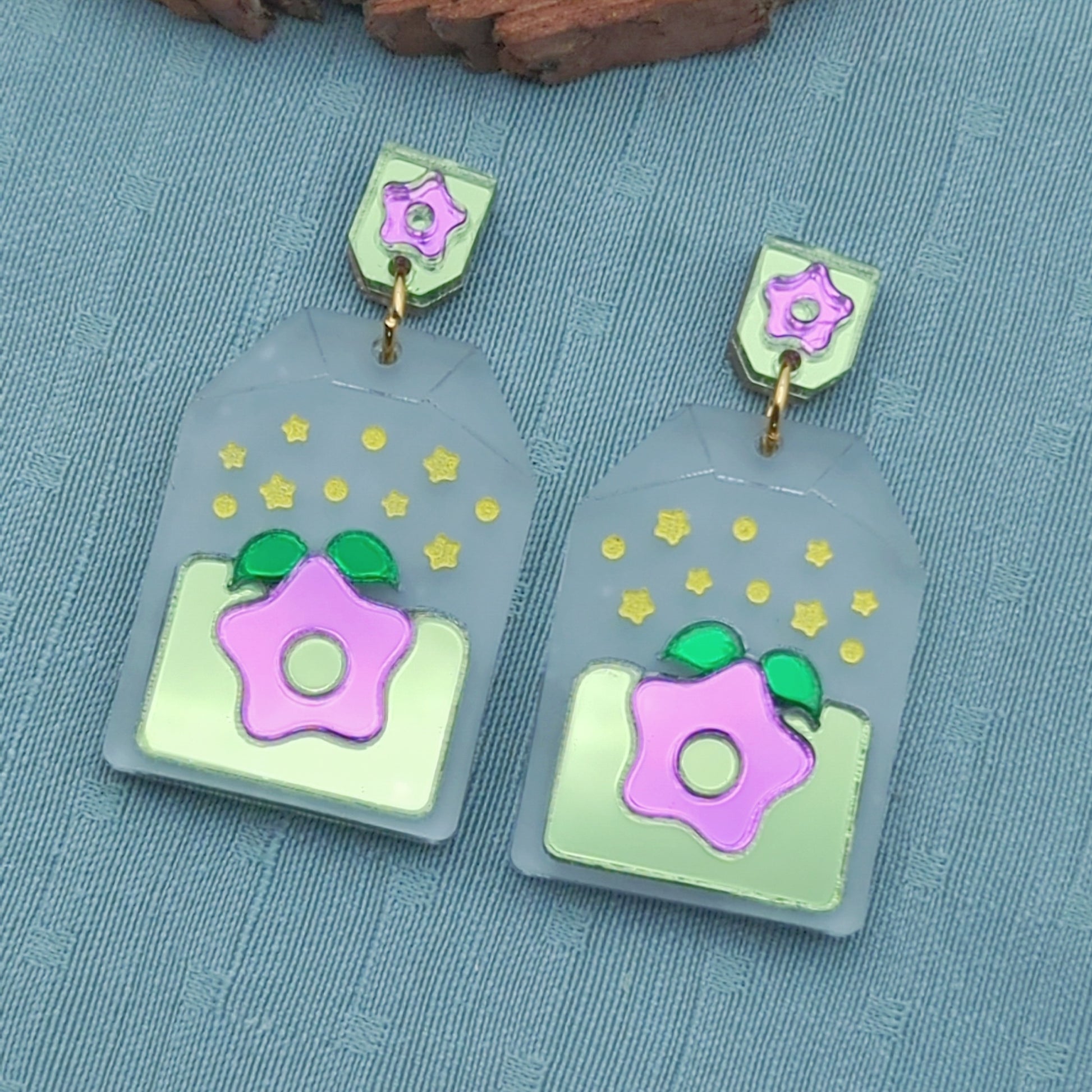 Frosted clear tea bag shaped earrings with a purple starfruit in the middle. The star has green leaves and is surrounded by light green. There are yellow star accents. Gold-toned jump rings connect to tag shaped stud posts that are green with a purple star.