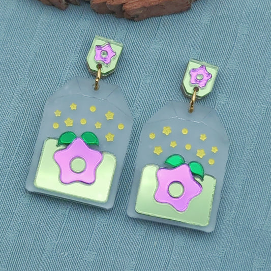 Frosted clear tea bag shaped earrings with a purple starfruit in the middle. The star has green leaves and is surrounded by light green. There are yellow star accents. Gold-toned jump rings connect to tag shaped stud posts that are green with a purple star.