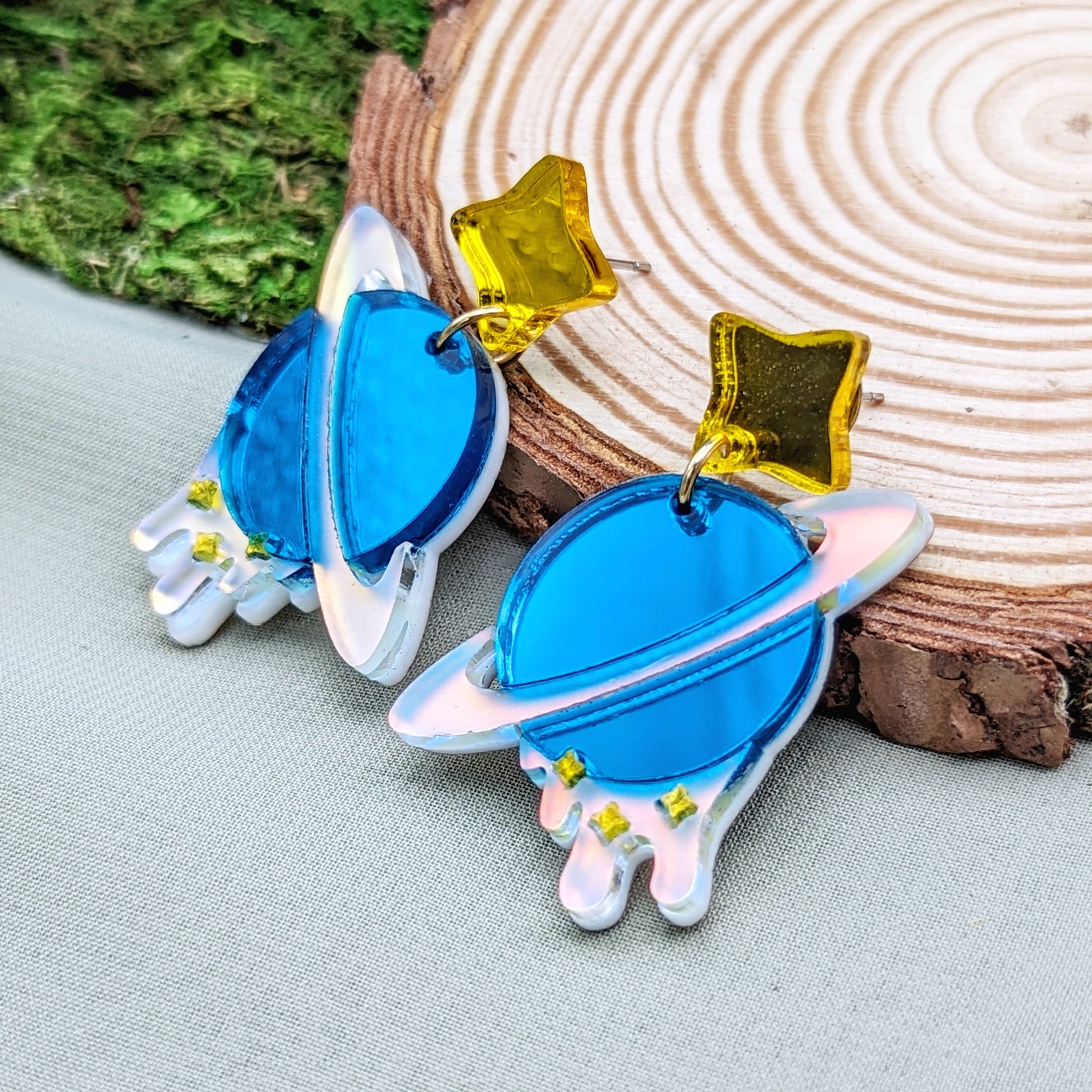 teal planet shaped dangle earrings with matte iridescent planetary rings, iridescent dripping below and small yellow stars with yellow star stud posts on a wood slice