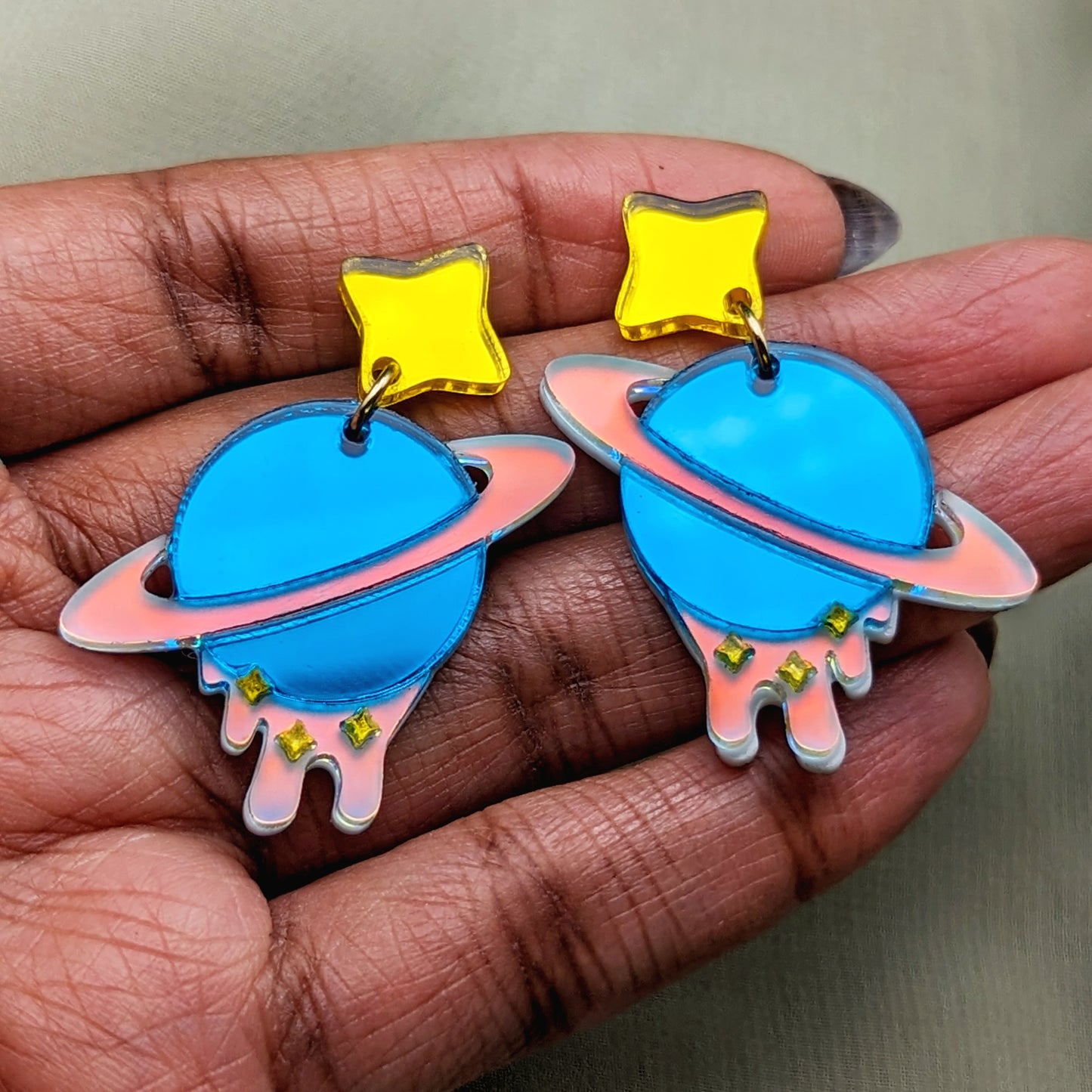 teal planet shaped dangle earrings with matte iridescent planetary rings, iridescent dripping below and small yellow stars with yellow star stud posts in hand for scale