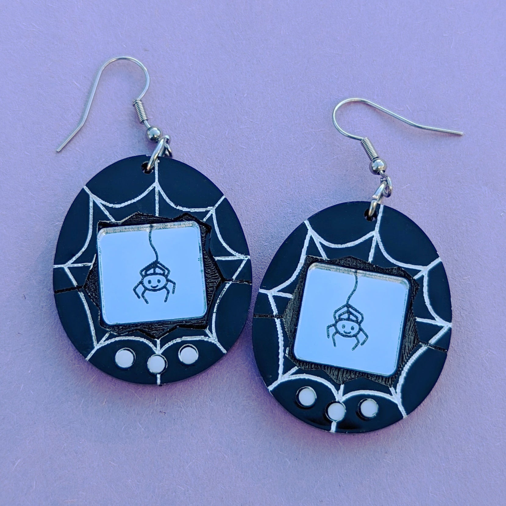 Halloween Spider Tamagotchi Acrylic Earrings with Stainless Steel Ear Hooks. Black and White earrings with a hand-painted white web, and a silver engraved screen featuring a cute spider hanging from it's web.