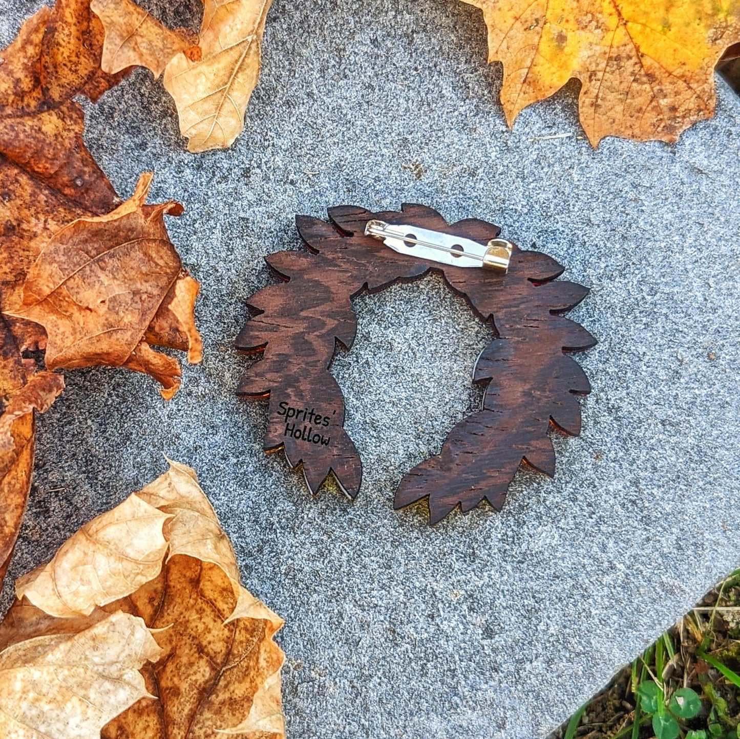 The back of a hades inspired laurel wreath brooch. There  is a 1 inch long pin across the top, glued to a dark wood backing with the name "Sprite's Hollow" engraved..